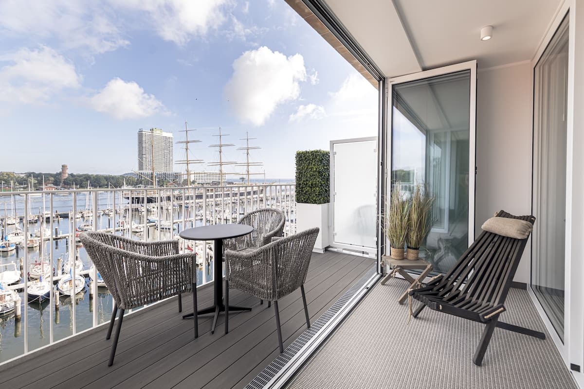 Een balkon met rieten stoelen en een tafel, met uitzicht op een jachthaven vol boten en grote zeilschepen, en een modern gebouw op de achtergrond.
