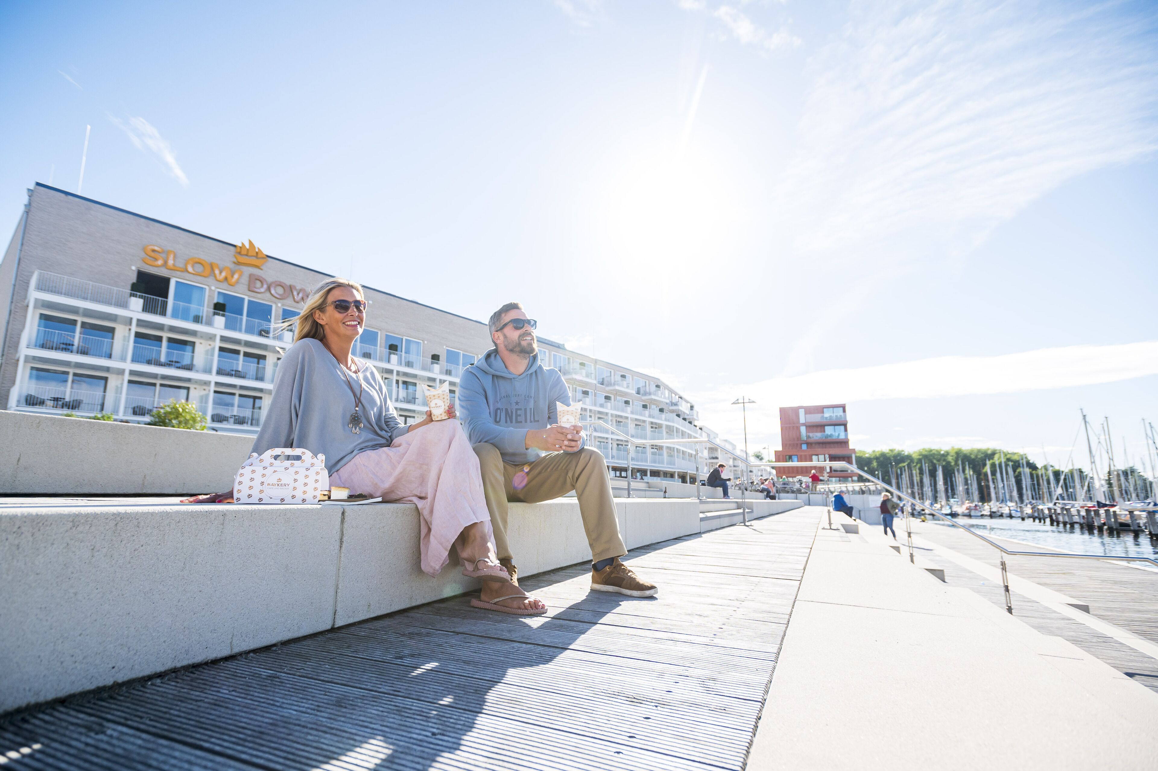 A couple sitting on a concrete ledge with a marina and boats in the background.