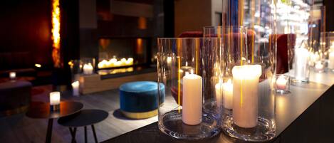 A bar area with candles in glass holders, a blue round seating, and a fireplace in the background.