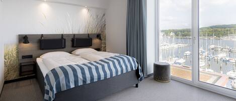 Habitació d'hotel moderna amb un llit gran, una tauleta de nit, una làmpada i vista a un port esportiu a través de grans finestres.