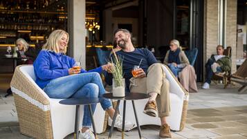 Dos personas sentadas en sillas de mimbre en un café al aire libre, con una mesa entre ellas, un bar al fondo y otros clientes a lo lejos.
