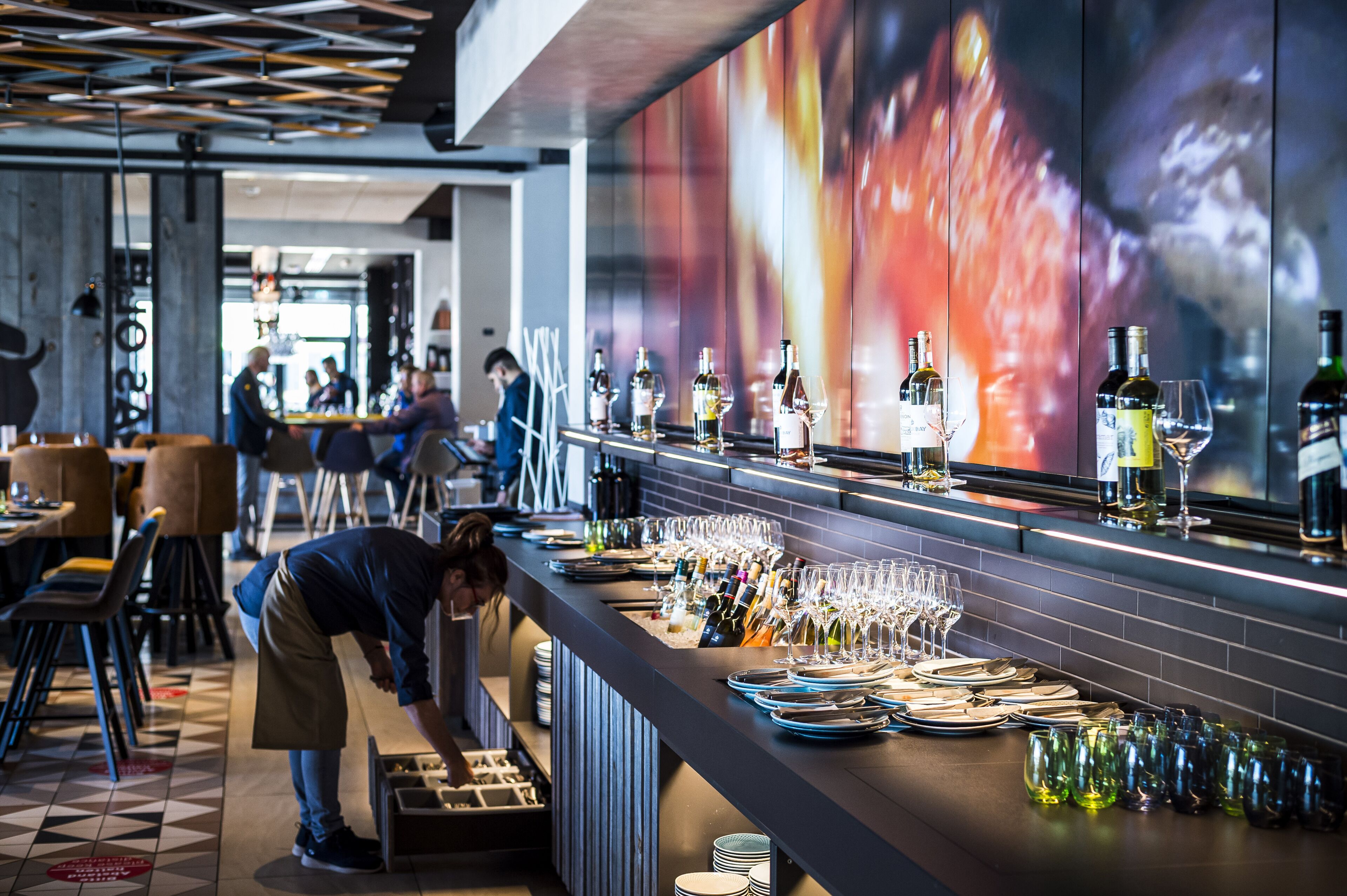 A bar area with bottles and glasses arranged on the counter, a person serving, and a modern interior with a geometric ceiling design.