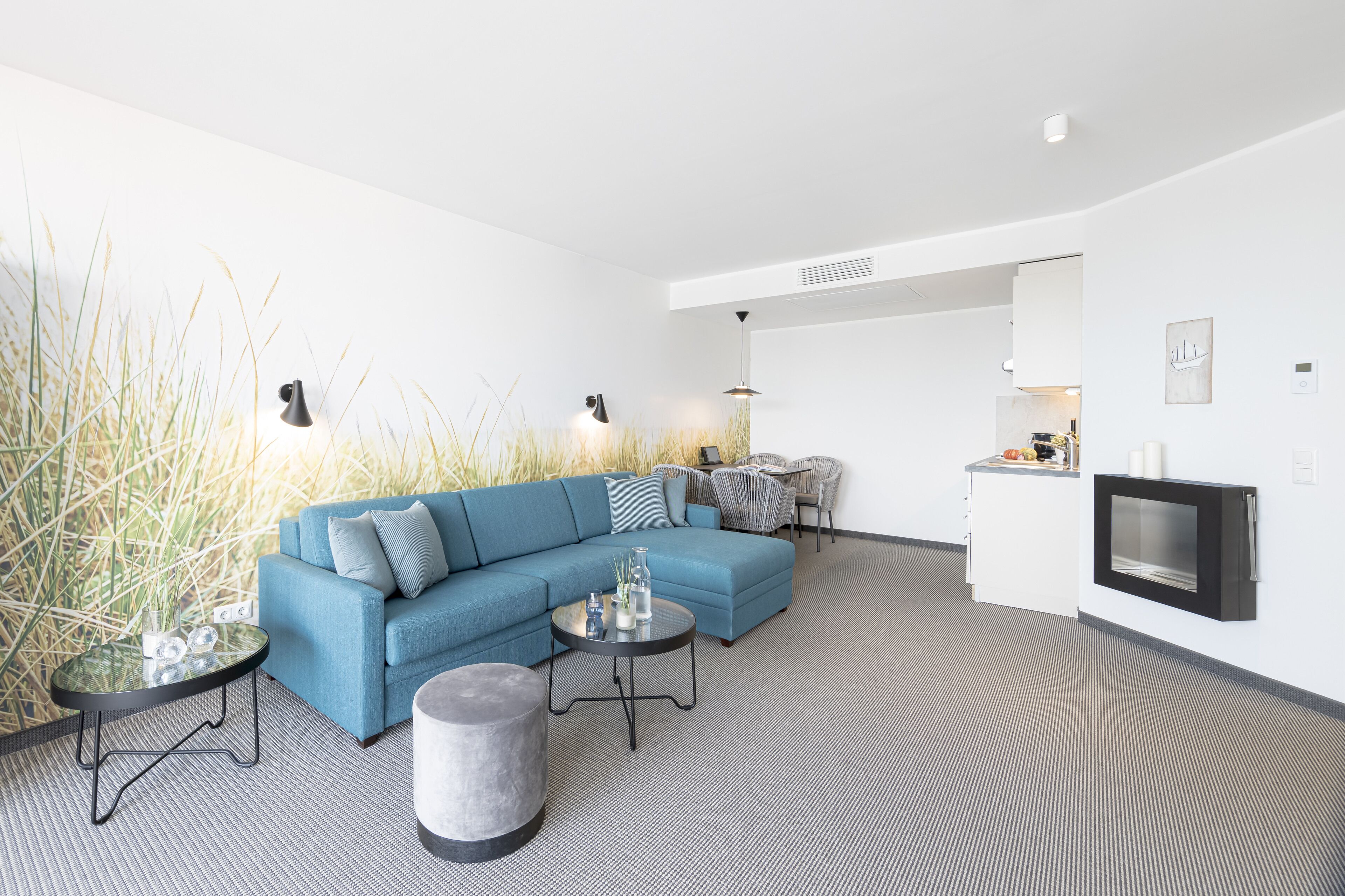 A modern living room with a blue sofa, two side tables, a round ottoman, and a kitchen area in the background.