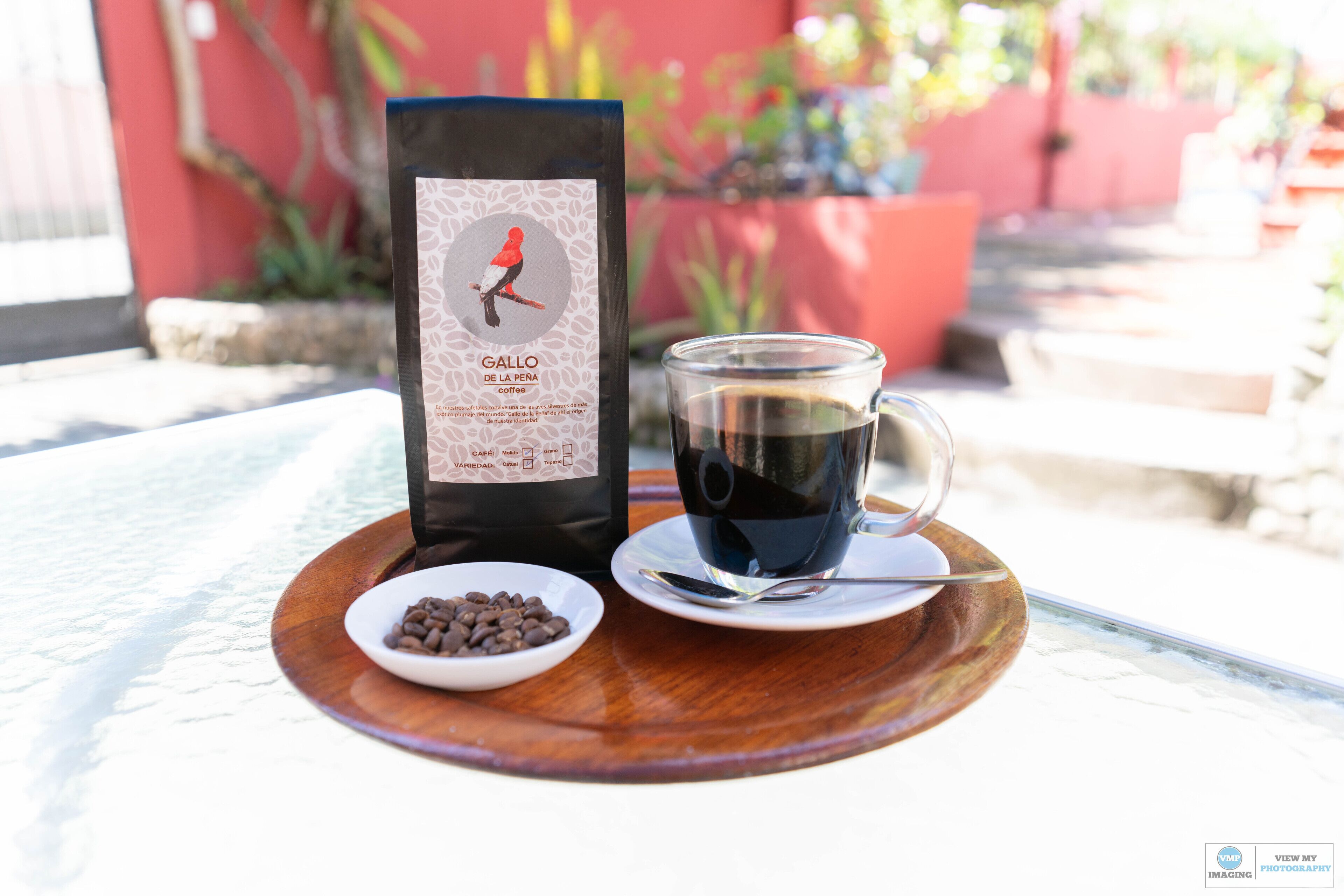 Una taza de café sobre un platito con una cuchara, un cuenco pequeño de granos de café y una bolsa de café en una bandeja de madera.