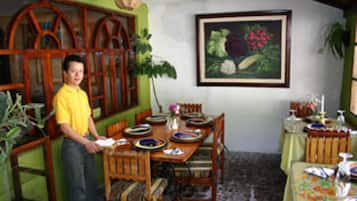 Uma sala de jantar com uma mesa de madeira posta para quatro pessoas, rodeada por cadeiras. Há uma pessoa de pé perto da mesa e um quadro na parede acima dela.