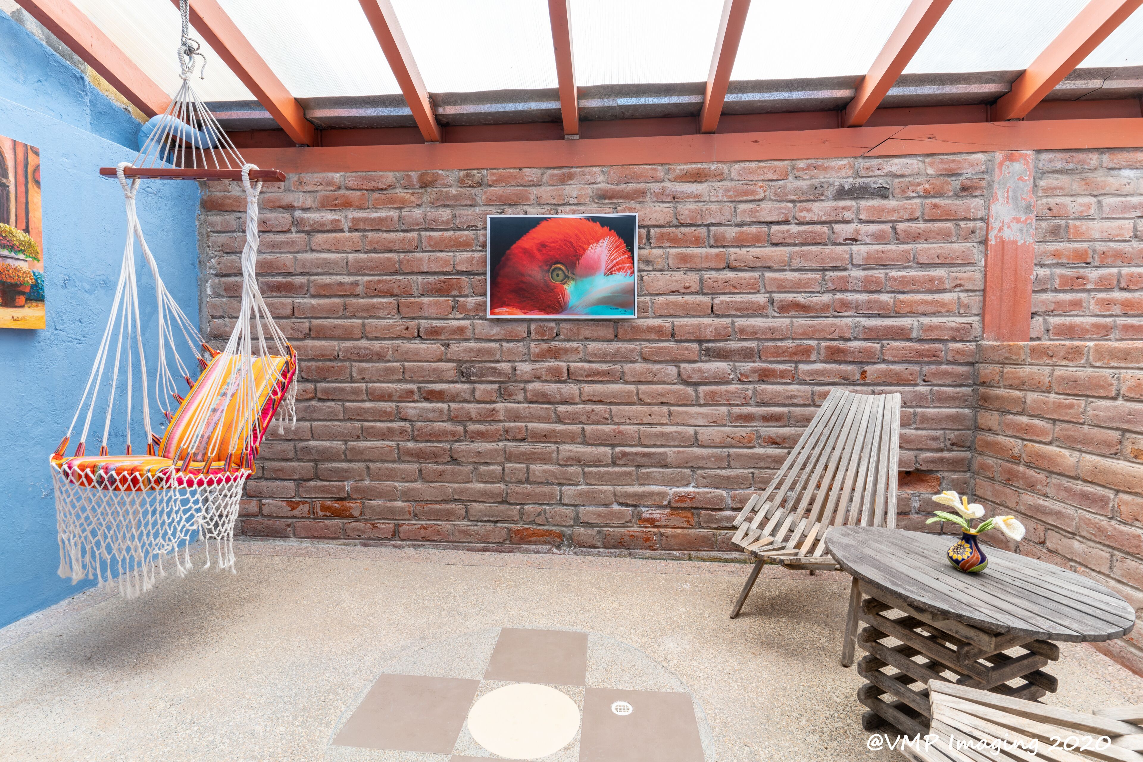 A patio area with a hammock, a wooden table with a vase, and a brick wall with a framed picture.