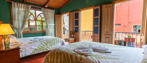 Quarto de hotel com duas camas, janela com cortinas, varanda com guarda-corpo e uma luminária.