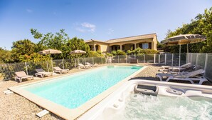 A swimming pool with lounge chairs and umbrellas, surrounded by a fence and a building in the background. - Villa L'Olivier with Private Pool and Spa in South Ardèche (Joyeuse)