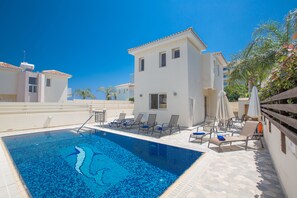 A swimming pool with lounge chairs and umbrellas, surrounded by a white building with a tiled roof. - Villa Petra, Modern 3BDR Protaras Villa with Pool, In The Centre of Protaras (Protaras)