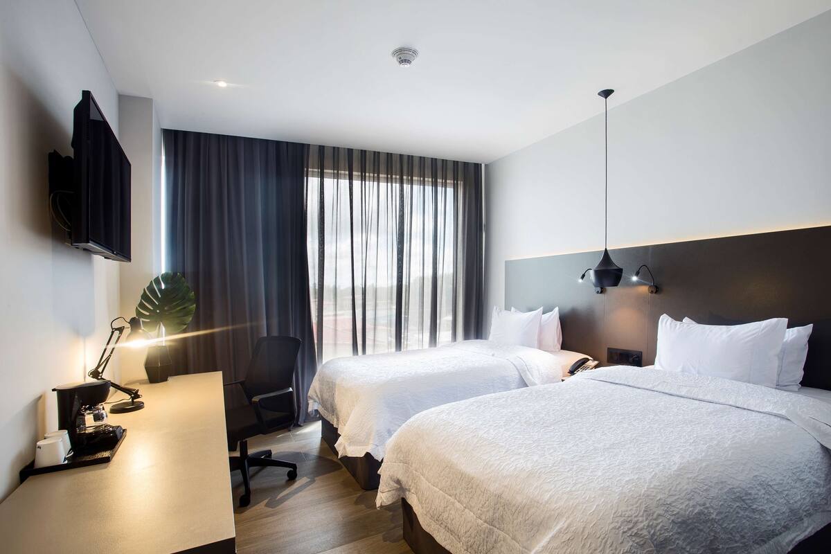 A hotel room with two beds, a desk with a lamp, a chair, a TV mounted on the wall, and a large window with curtains.