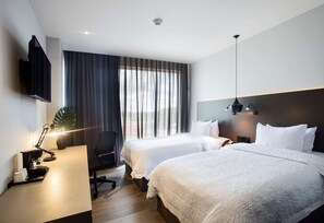Une chambre d’hôtel avec deux lits, un bureau avec une lampe, une chaise, un téléviseur fixé au mur et une grande fenêtre avec des rideaux.