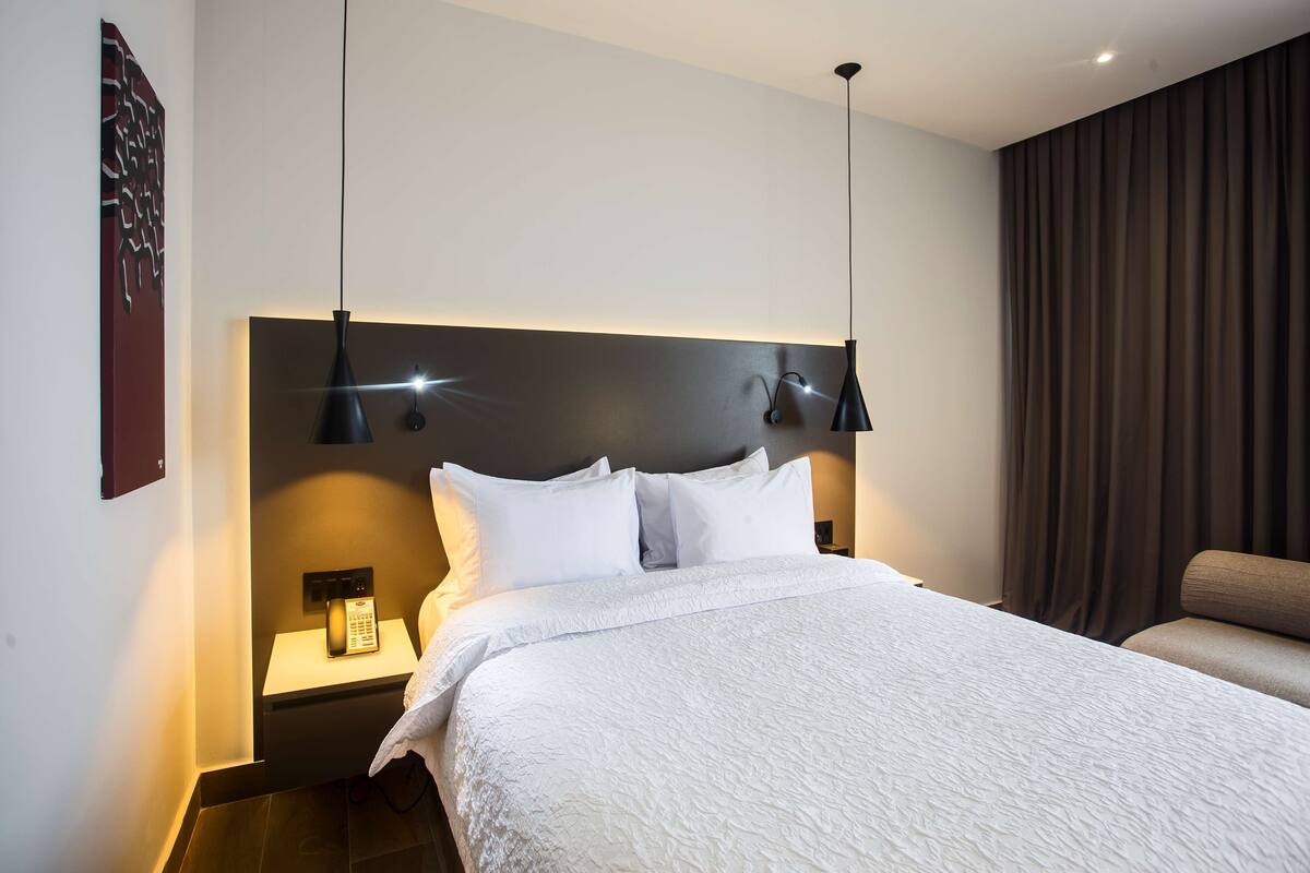 A hotel room with a large bed, two bedside lamps, a nightstand with a phone, and a dark curtain.