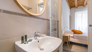 A bathroom with a white sink, mirror, and towel rack. There is a window with curtains and a bed with an orange pillow in the adjacent room.