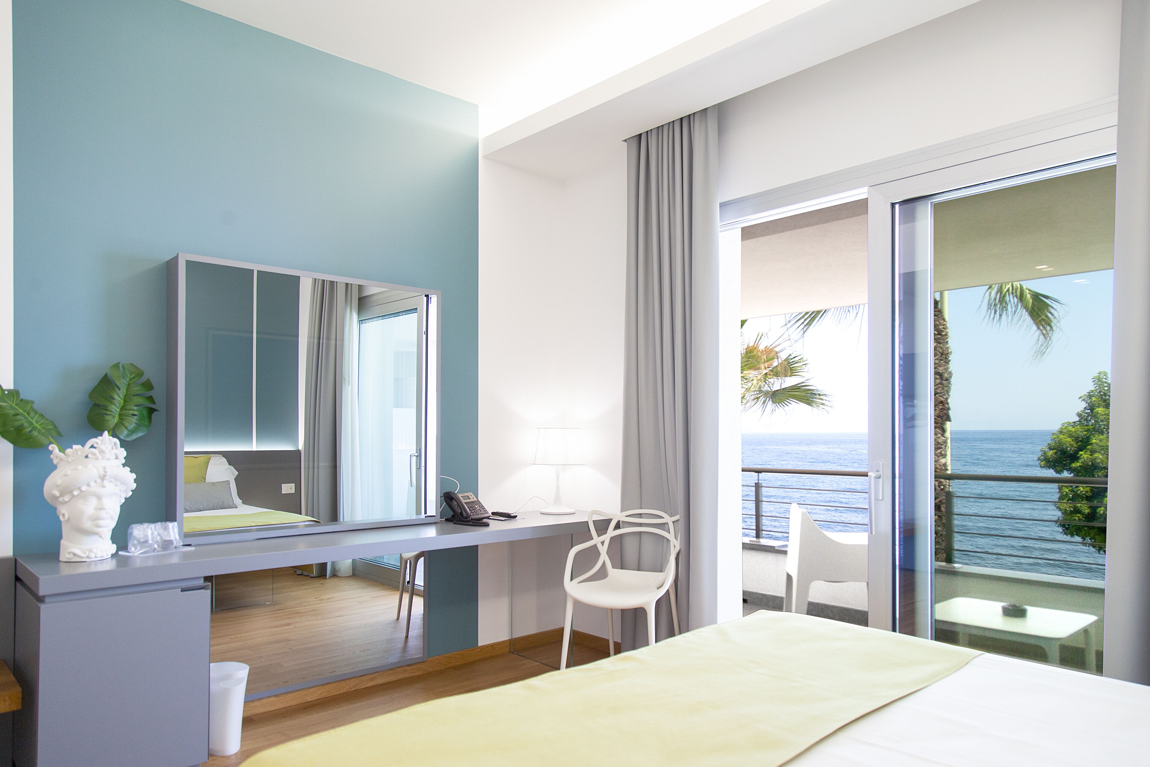 Une chambre d’hôtel moderne dotée d’un grand lit, d’un bureau avec une chaise, d’une petite table, d’un miroir et d’un balcon offrant une vue sur la mer.