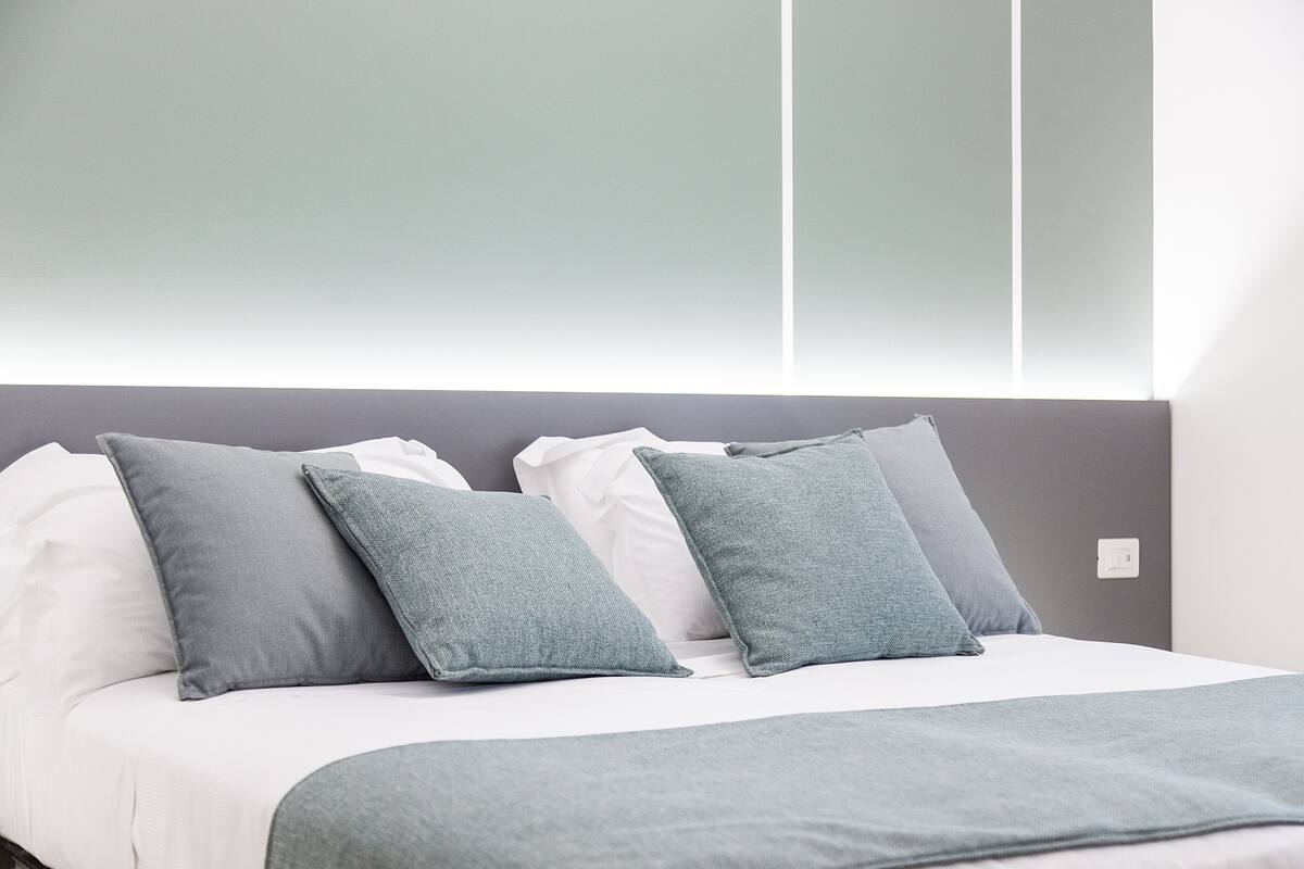 Un lit bien fait, avec une literie blanche et grise, une tête de lit grise et un plaid gris.