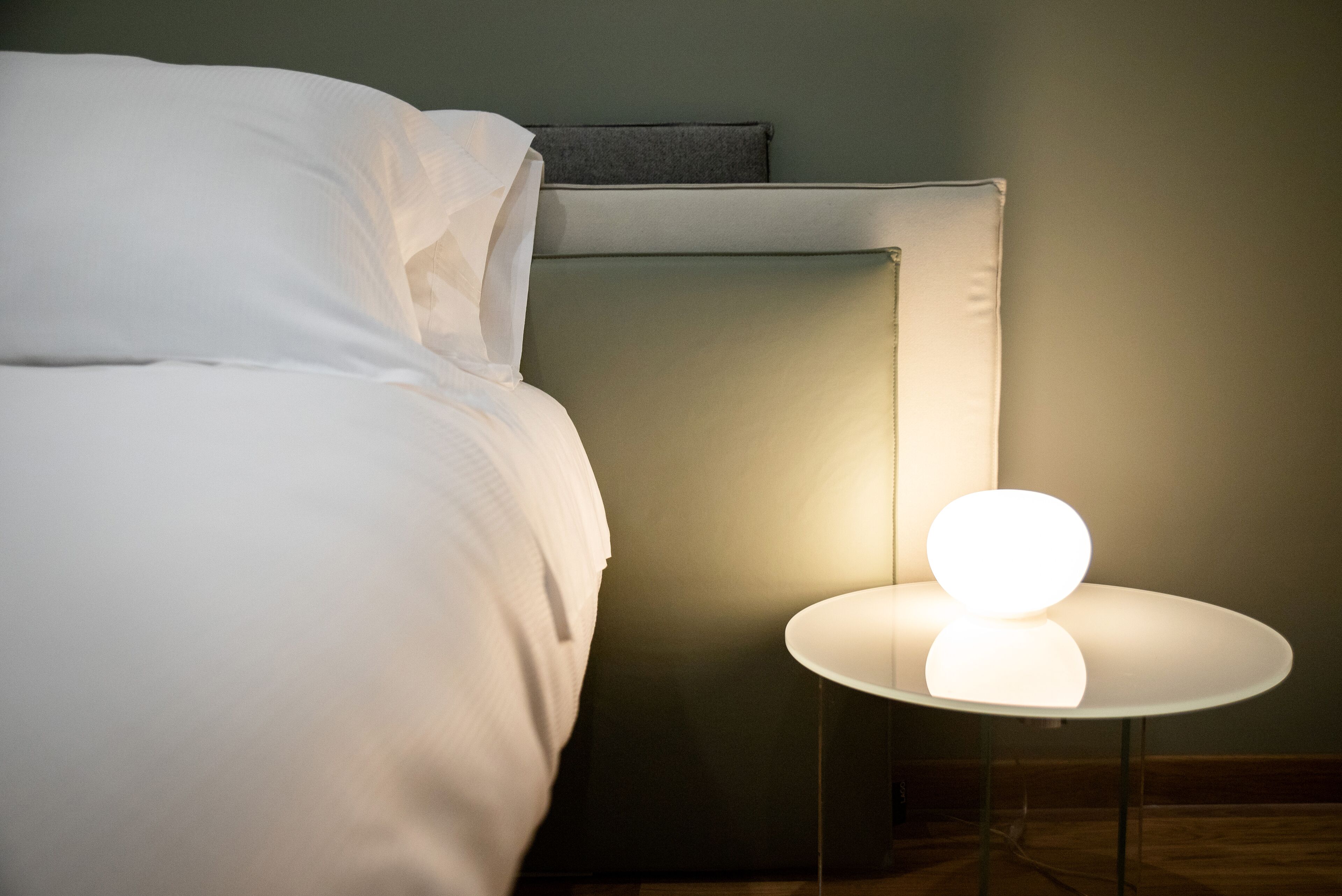 Una cama bien hecha con sábanas blancas, una mesita de noche con una lámpara redonda y un cabecero oscuro.