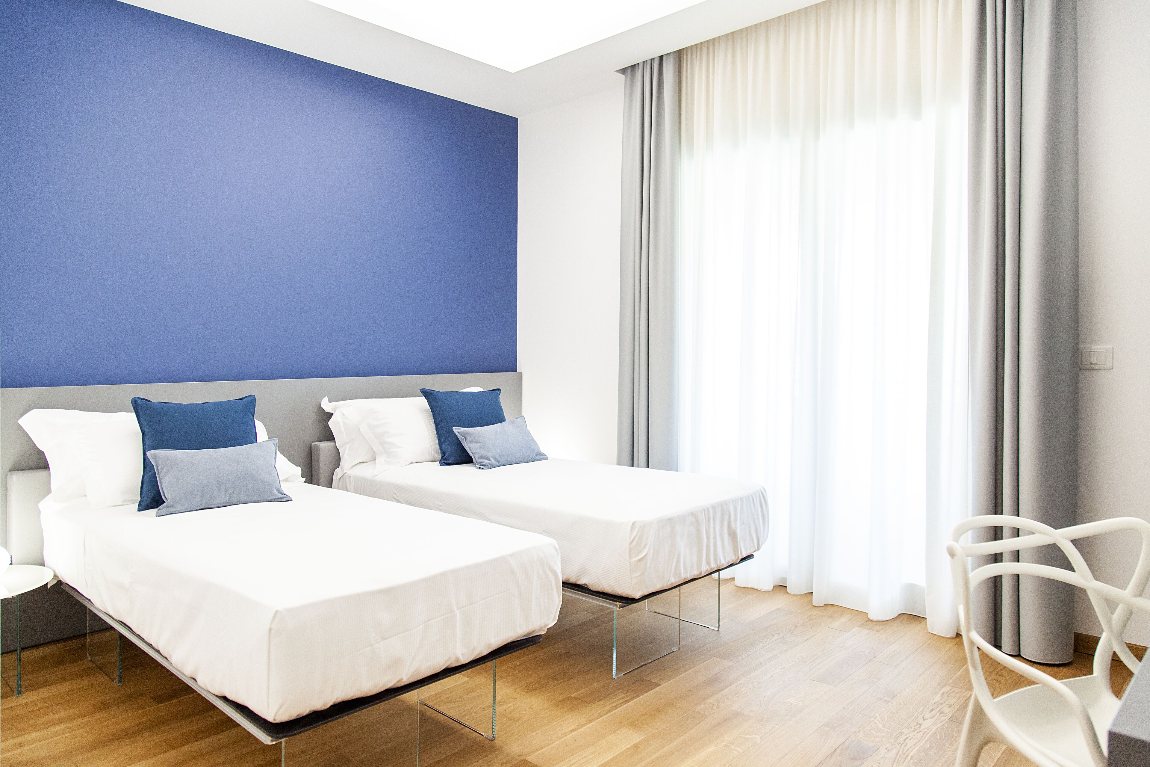 Una camera d'albergo con due letti, una sedia bianca e un'ampia finestra con tende.