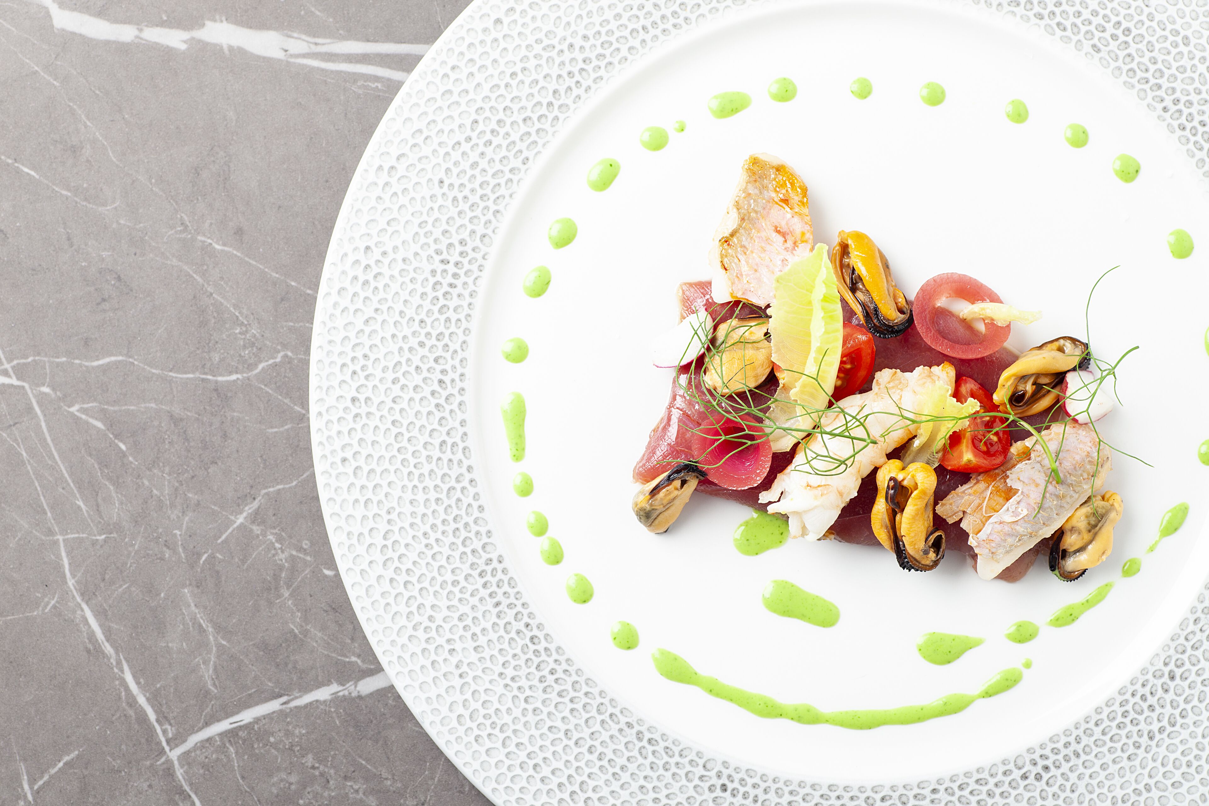 A white plate with a seafood dish, garnished with green sauce and herbs, on a marble surface.