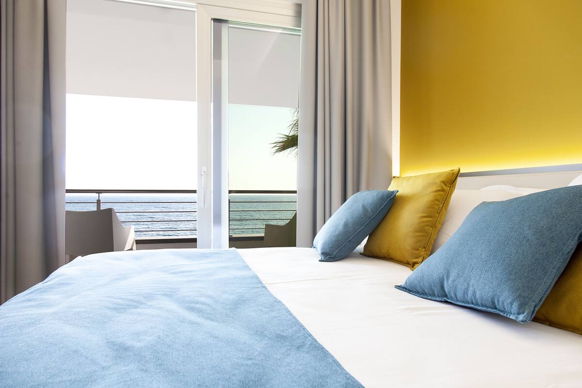 Une chambre d’hôtel avec un lit, une vue sur la mer grâce à une porte coulissante et un mur d’accent jaune.
