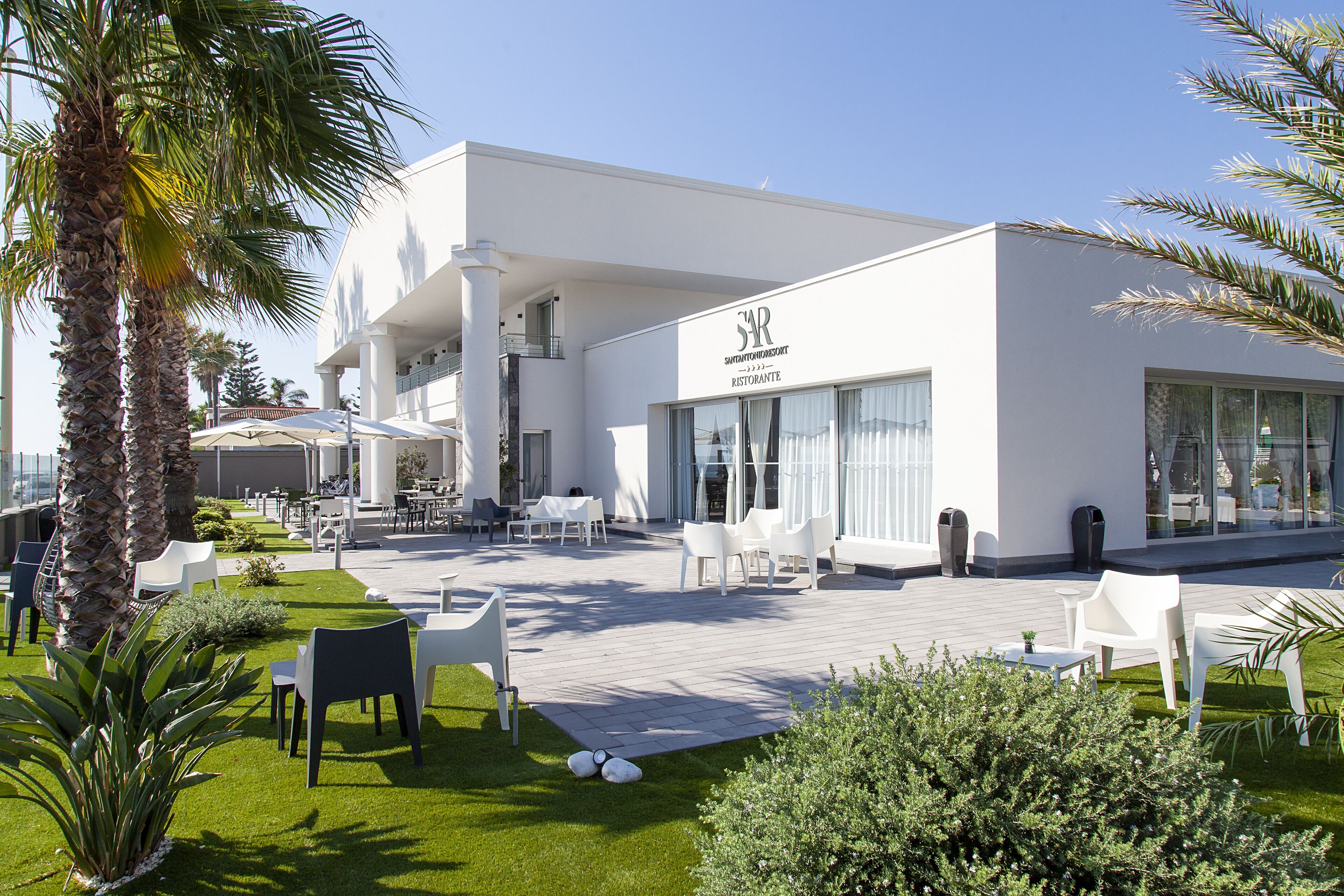 Un bâtiment moderne à l’extérieur blanc, doté d’un espace extérieur abrité avec des chaises et des tables blanches, et d’un jardin bien entretenu avec des palmiers et des arbustes.