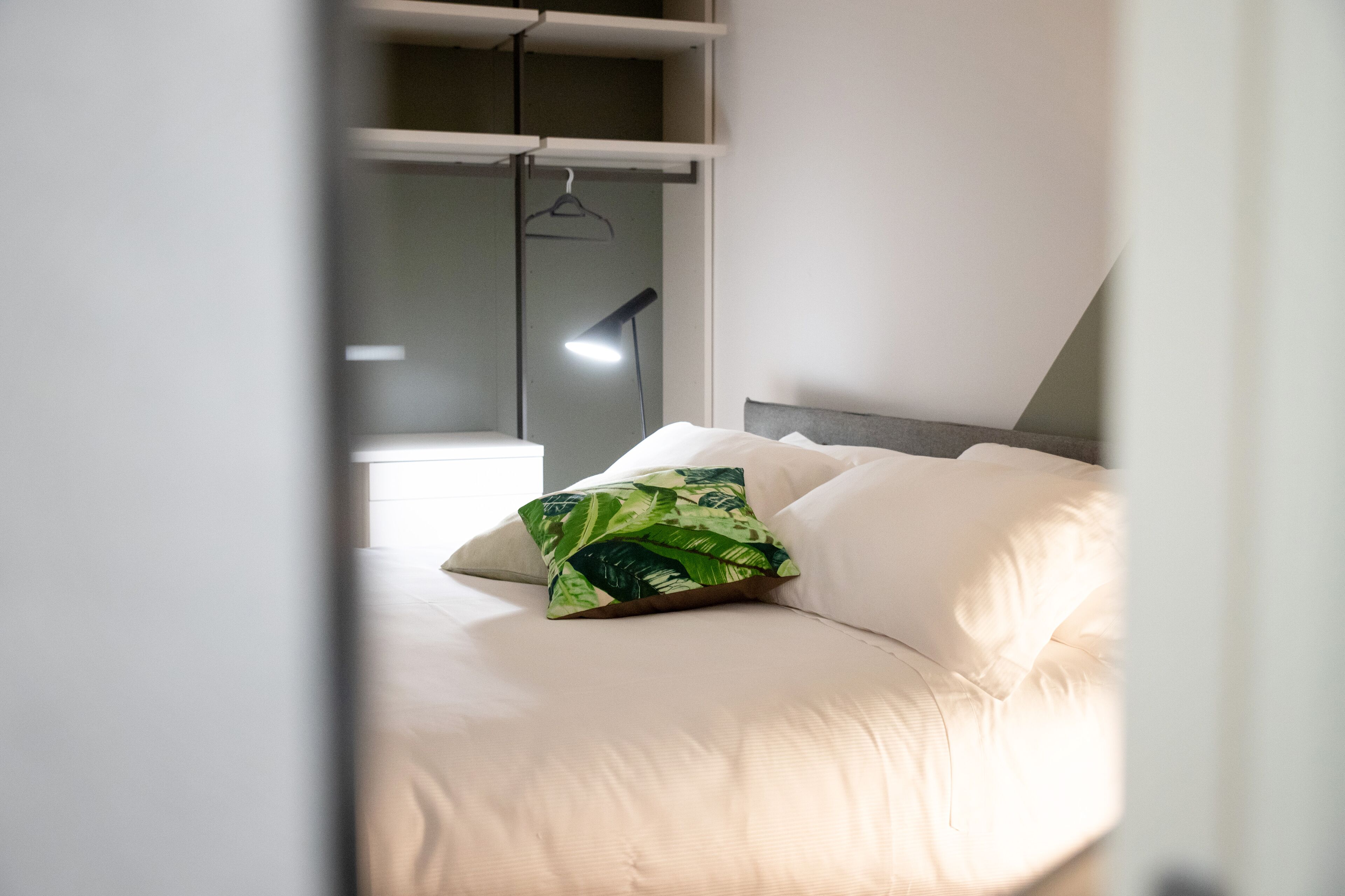Cama bem arrumada com lençóis brancos e uma almofada estampada com padrão de folhagem verde.