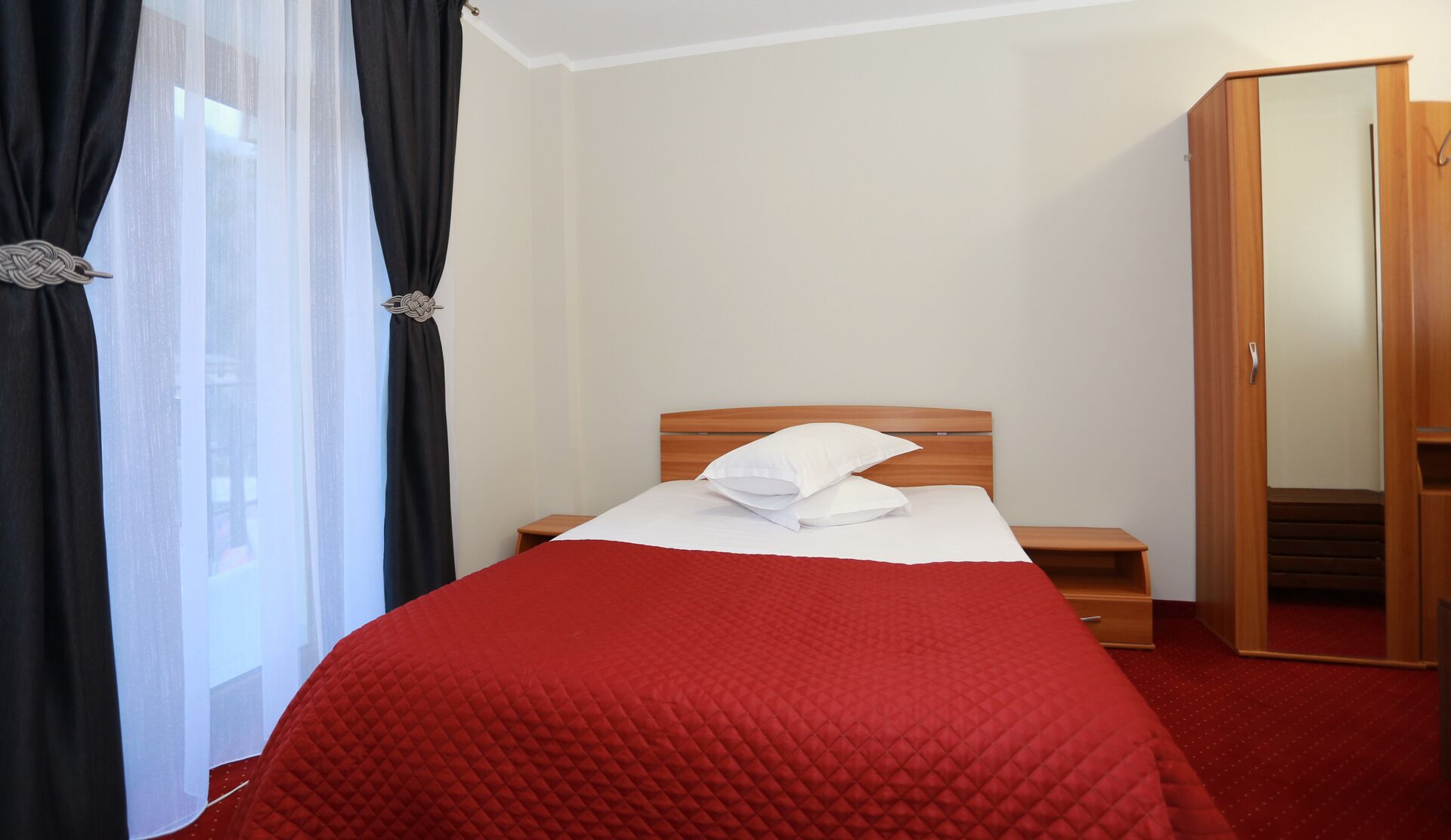 Una habitación de hotel con una cama, una mesita de noche, un armario de madera y una ventana con cortinas negras.