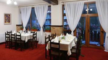 Comedor con mesas dispuestas para dos, cortinas blancas y vistas a edificios a través de grandes ventanales.