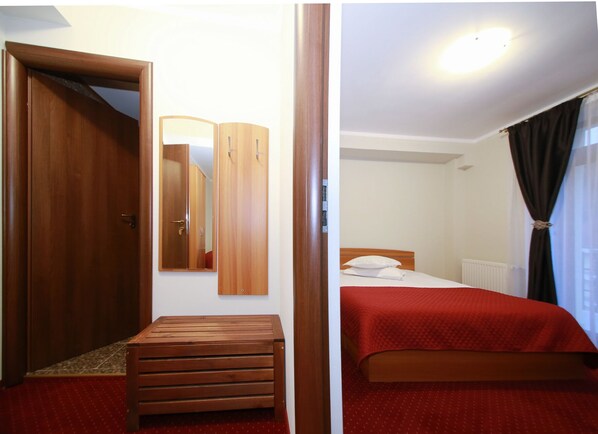 Una habitación con un armario de madera, un banco, una cama con colcha roja y un espejo en la pared.