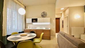 A modern kitchen with a central island, a dining area with a round table and chairs, a sofa, and a clock on the wall. - Velvet 1 (Verona)