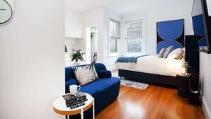 Une chambre moderne avec un lit, un canapé bleu, une petite table ronde avec des livres et une tasse, et une grande fenêtre avec des stores.