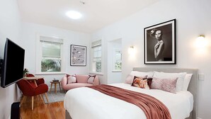 A modern bedroom with a large bed, a red chair, a small table, and artwork on the walls. - Perfect Located Cosy Studio in Sydney (Lewisham)