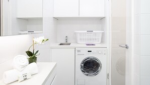 A modern, white-walled laundry room with a front-loading washing machine, a drying rack, and a small sink. - Perfect Located Cosy Studio in Sydney (Lewisham)