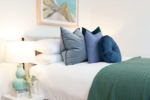 A neatly made bed with a white duvet and multiple pillows in shades of blue and gray. A green textured throw blanket is draped over the bed. A teal lamp with a white shade is on a bedside table beside the bed, which also holds a small vase with flowers and a book titled "Tana French". - Art Deco Designer Studio in Lewisham (Lewisham)