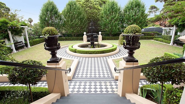 Un escalier menant à un jardin agrémenté d’une fontaine, le tout bordé de haies et d’arbres taillés avec soin.