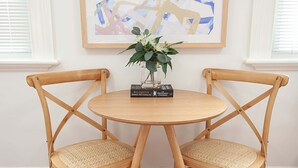 Une table ronde en bois, accompagnée de deux chaises en osier, un vase rempli de fleurs et, au mur, un tableau abstrait encadré.