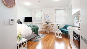 Une chambre moderne et bien éclairée, équipée d’un lit, d’un bureau, d’une chaise, d’un téléviseur et d’une horloge murale.