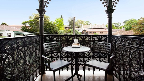 A balcony with ornate metal railings and two chairs, a small round table with a book and a cup, and a view of houses and trees in the background. - Beautiful Studio in Heritage Building with Balcony (Lewsiham)
