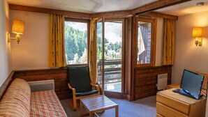 Une pièce confortable avec un lit, une chaise, une petite table, un bureau avec un ordinateur et de grandes fenêtres donnant sur l’extérieur.
