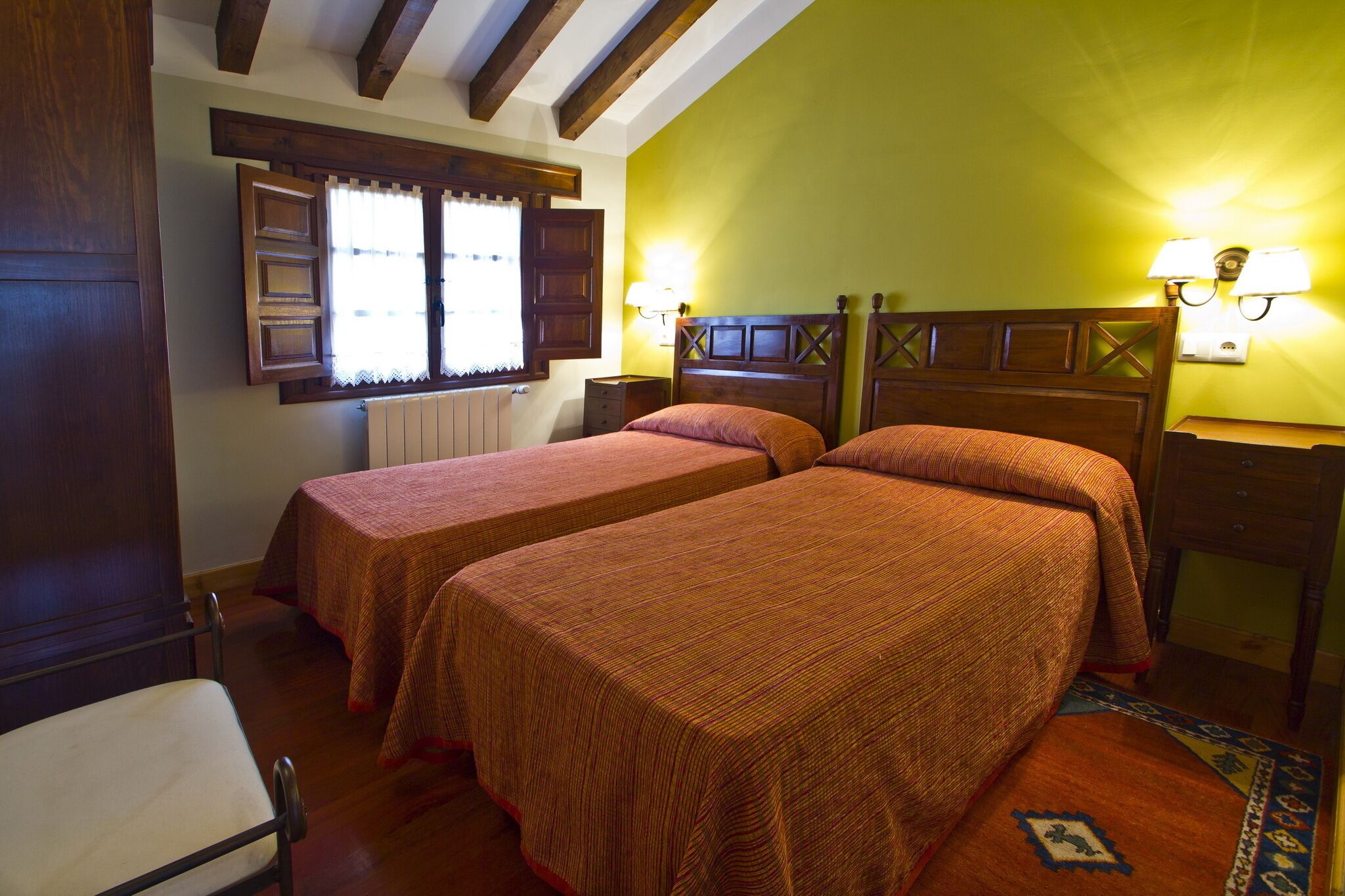 A room with two single beds, wooden headboards, a window with curtains, a nightstand with lamps, and a wooden floor.