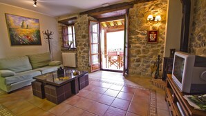 Un accogliente soggiorno con divano, tavolino e televisione. Presenta pareti in pietra, soffitto in legno e vista su un patio esterno attraverso una porta in vetro.