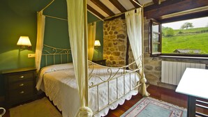 Un letto a baldacchino con una parete in pietra sullo sfondo, due lampade ai comodini e vista su un paesaggio verde attraverso una finestra.