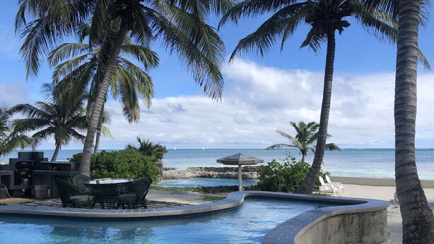 Área de piscina com espreguiçadeiras e mesa, cercada por palmeiras e vista para o mar.