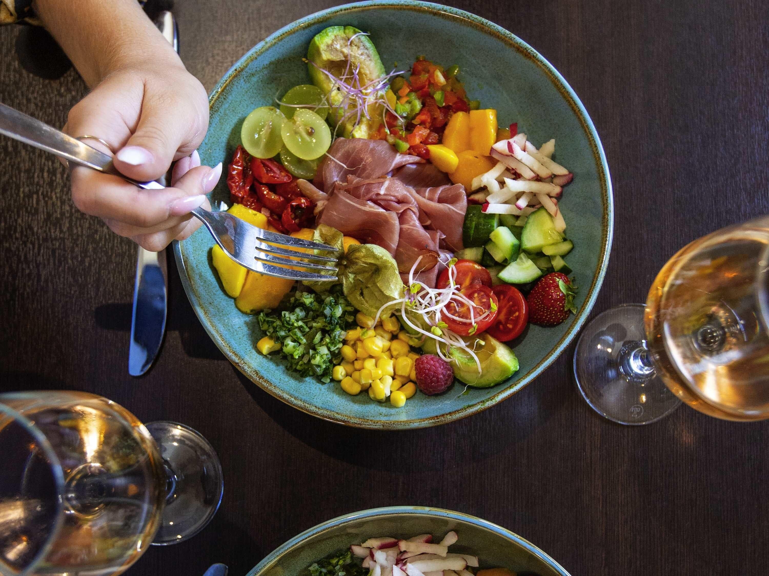 Uma tigela com frutas e legumes variados, um garfo e dois copos de vinho sobre uma mesa.