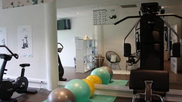 Ein Fitnessraum mit Trainingsgeräten, darunter Fahrrädern und einer Kraftmaschine, sowie Übungsbällen auf Matten.