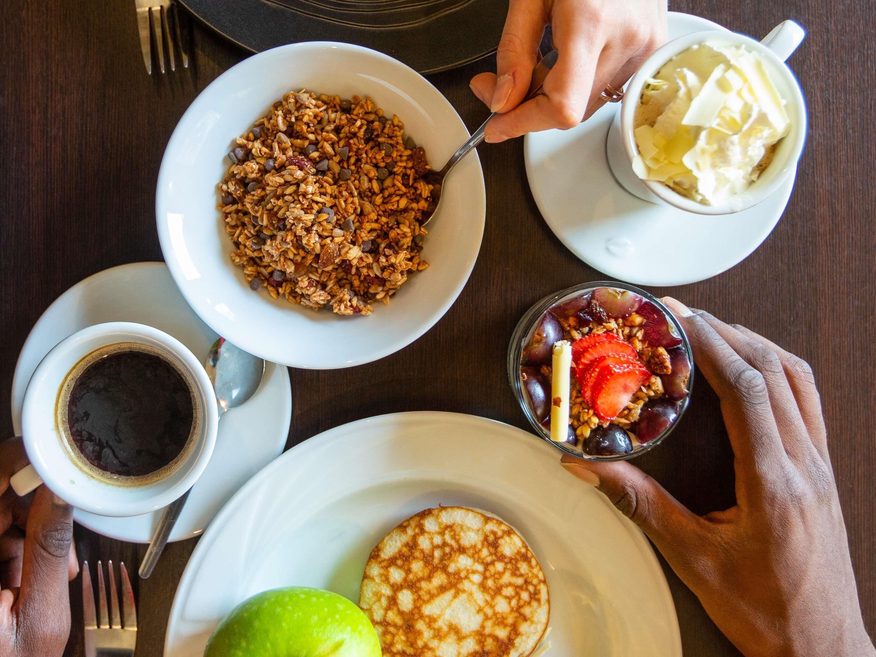 A breakfast setting with a bowl of granola, a cup of coffee, a glass of fruit, and a plate with pancakes.