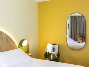 Habitació d'hotel amb un llit, una làmpada de nit, un mirall i una persona llegint un llibre.