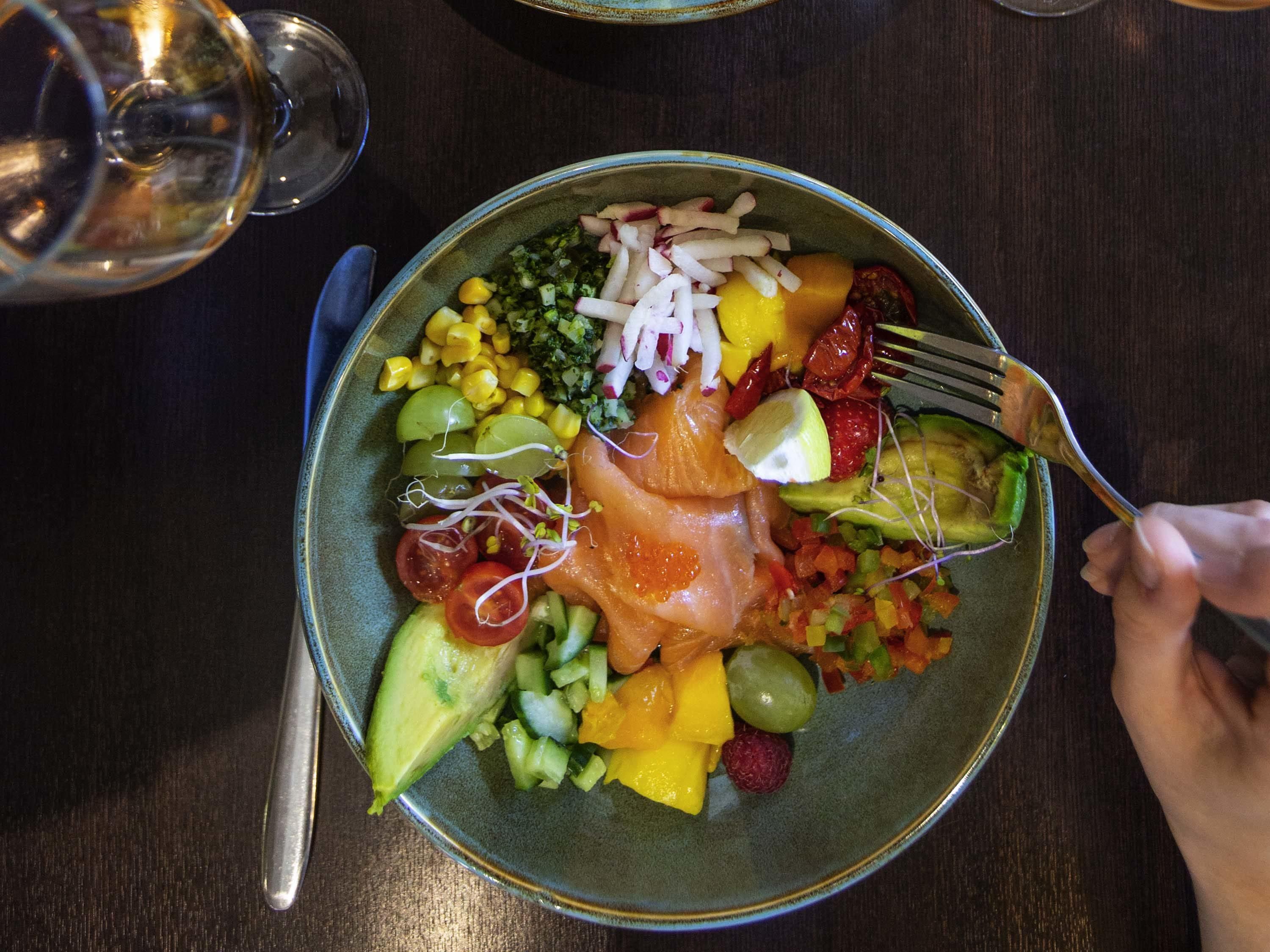 A plate of food with various fruits, vegetables, and salmon, accompanied by a fork and a glass of water.