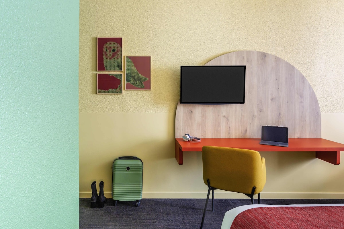 A hotel room with a bed, a desk with a TV, a suitcase, and artwork on the wall.