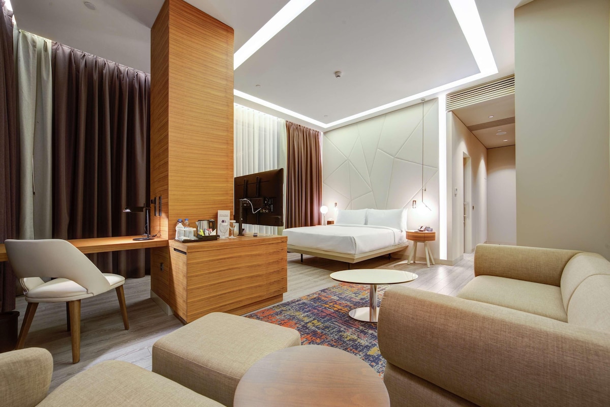 A modern hotel room with a bed, a desk, a chair, a sofa, and a small round table.