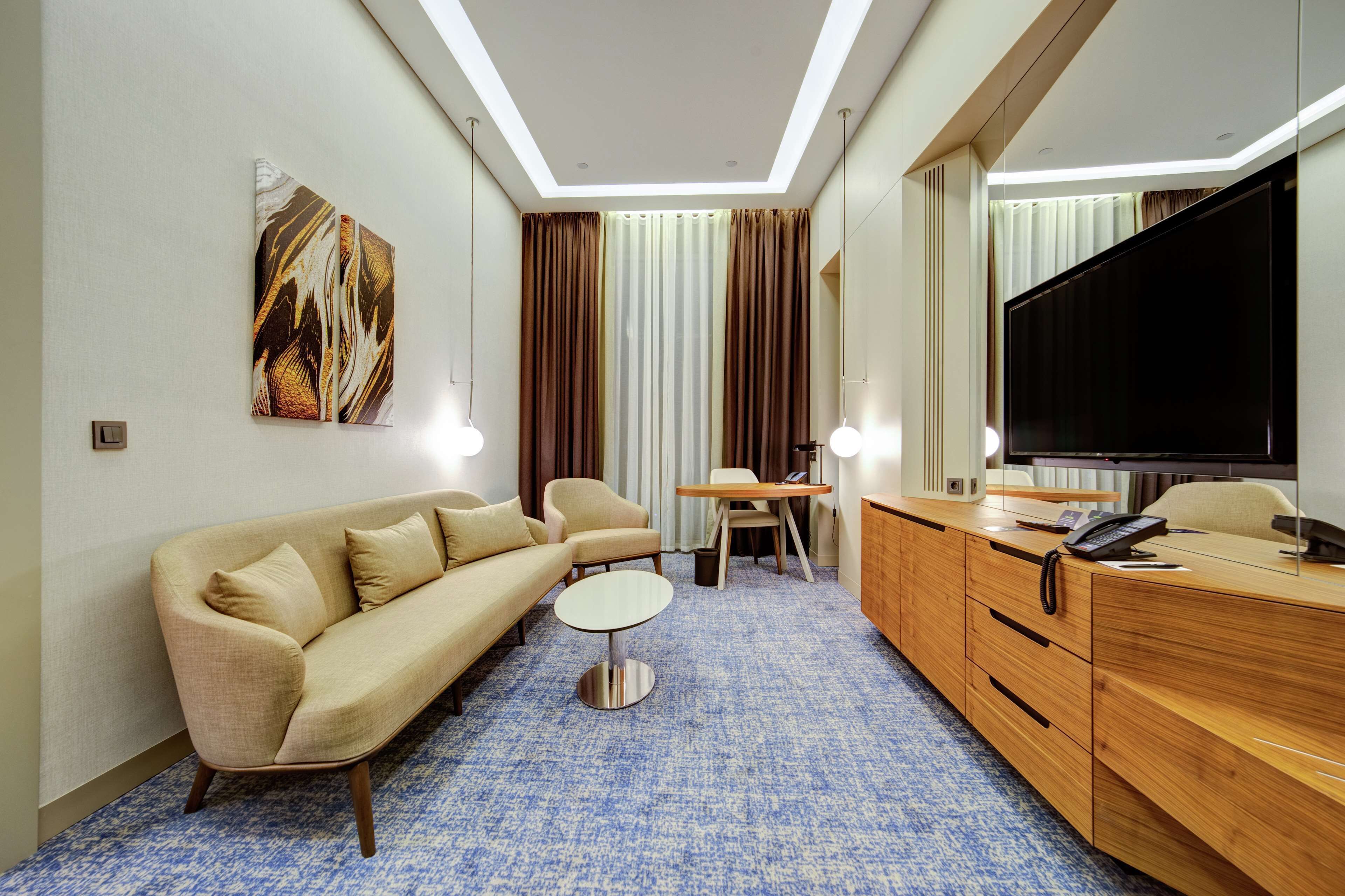 A modern hotel room with a sofa, a small round table, a wooden desk with a TV, and a large window with curtains.